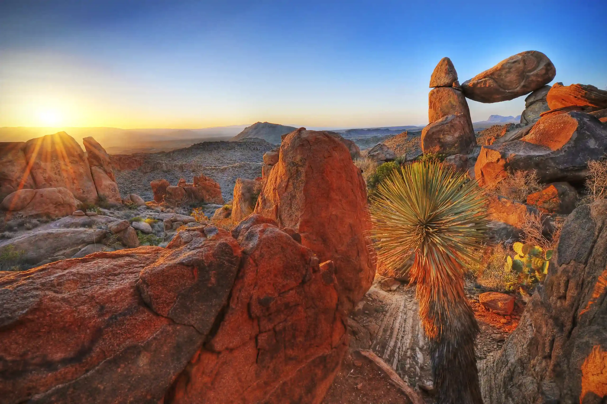 Red rocks balancing and desert plants in the foreground with a view of the Big Bend State Park with early rising sun and blue sky.