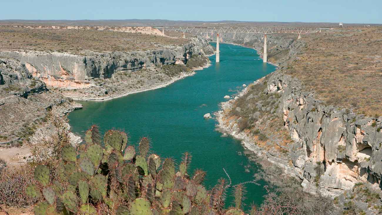 The Pecos River running through a canyon with the "High Bridge" suspension bridge in the background, and desert vegetation and cactus in the foreground.